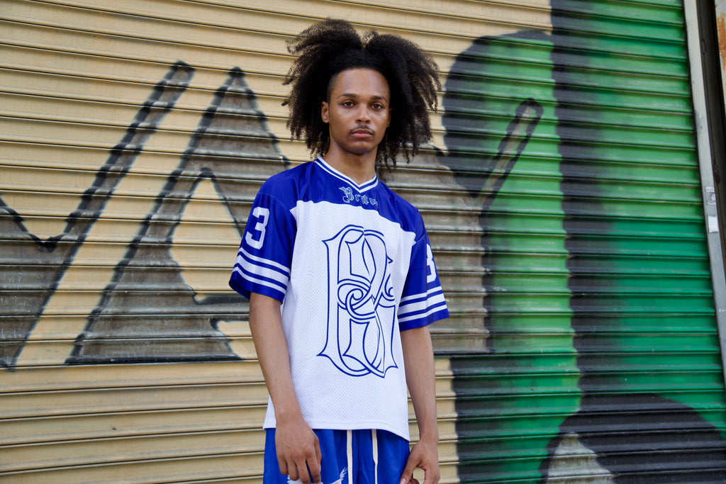 Person wearing a blue and white sports jersey with a logo, standing in front of a graffiti-covered wall.