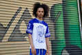 Person wearing a blue and white sports jersey with a logo, standing in front of a graffiti-covered wall.